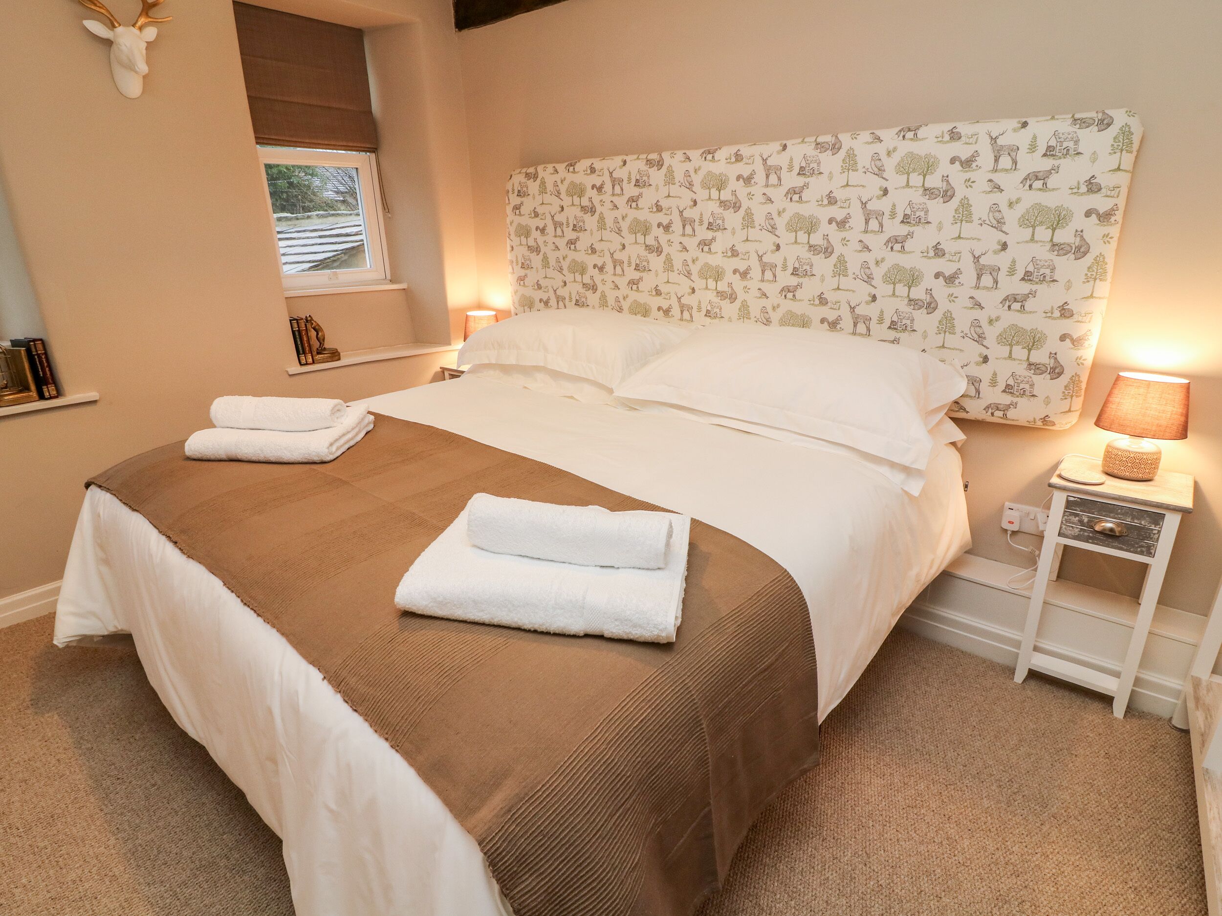 A neatly made bed with white and brown linens, a headboard with a nature-themed pattern, and a bedside table with a lamp.