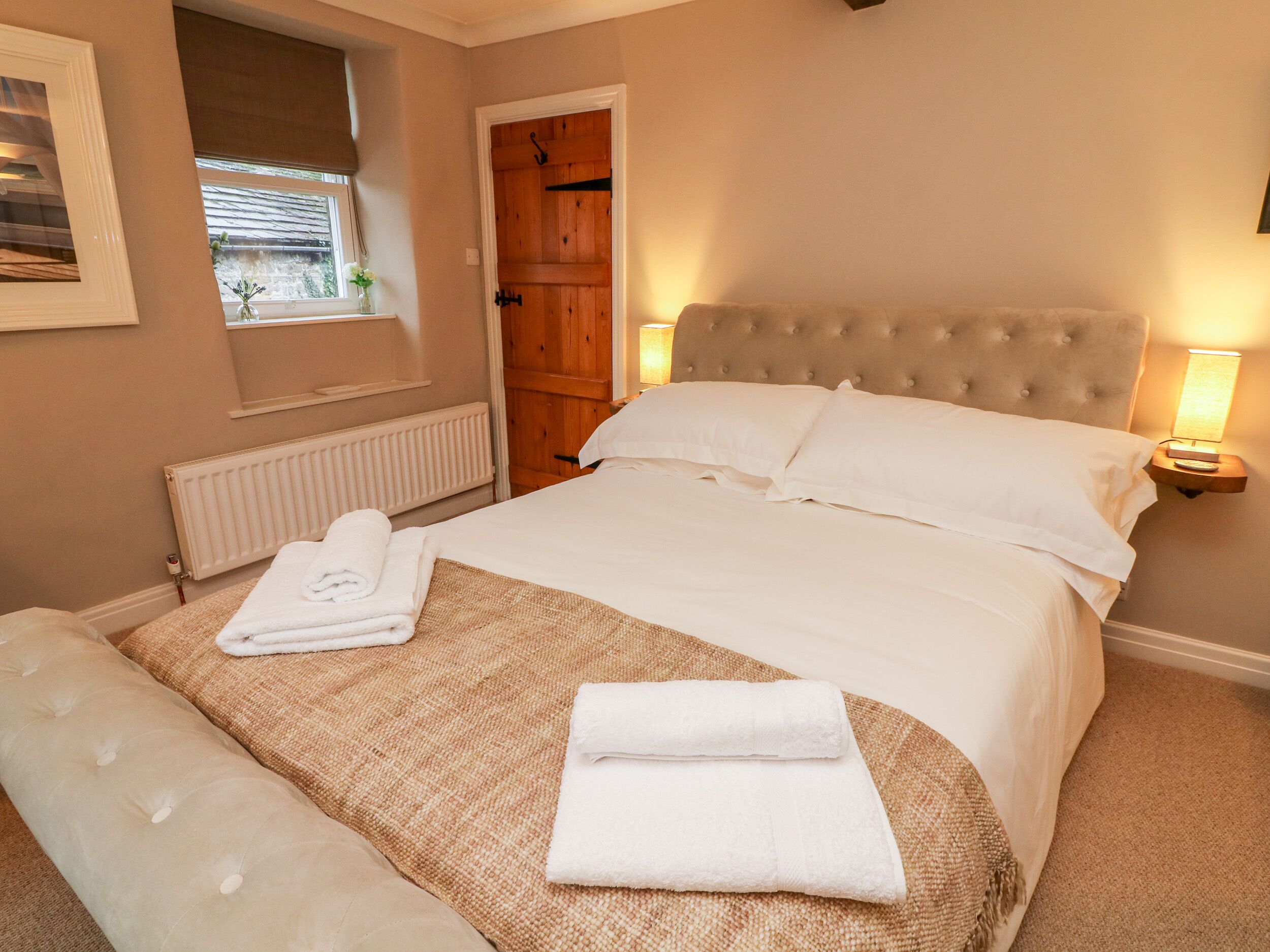 A neatly made bed with white linens and a headboard, a bedside lamp, a window with a view, and a radiator.