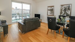 Una sala de estar moderna con un sofá gris, sillas negras, una mesa de comedor de vidrio y un amplio ventanal con vista a un puerto deportivo.