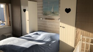 Une chambre à coucher avec un lit, une fenêtre, un tableau représentant un voilier et deux portes ornées de découpes en forme de cœur.