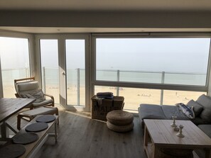 A room with a large window overlooking a beach, a sofa, a table, and a chair. - Seafront, 95 m2 5th floor apartment, 3 bedrooms, Wi-fi, Garage, Terrace (Koksijde)