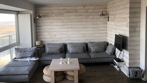 Un salon moderne avec un canapé d’angle gris, une petite table basse en bois et une grande fenêtre donnant sur la plage.