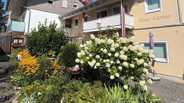 A building with a sign reading "Haus Carina" and a garden with various plants and flowers.