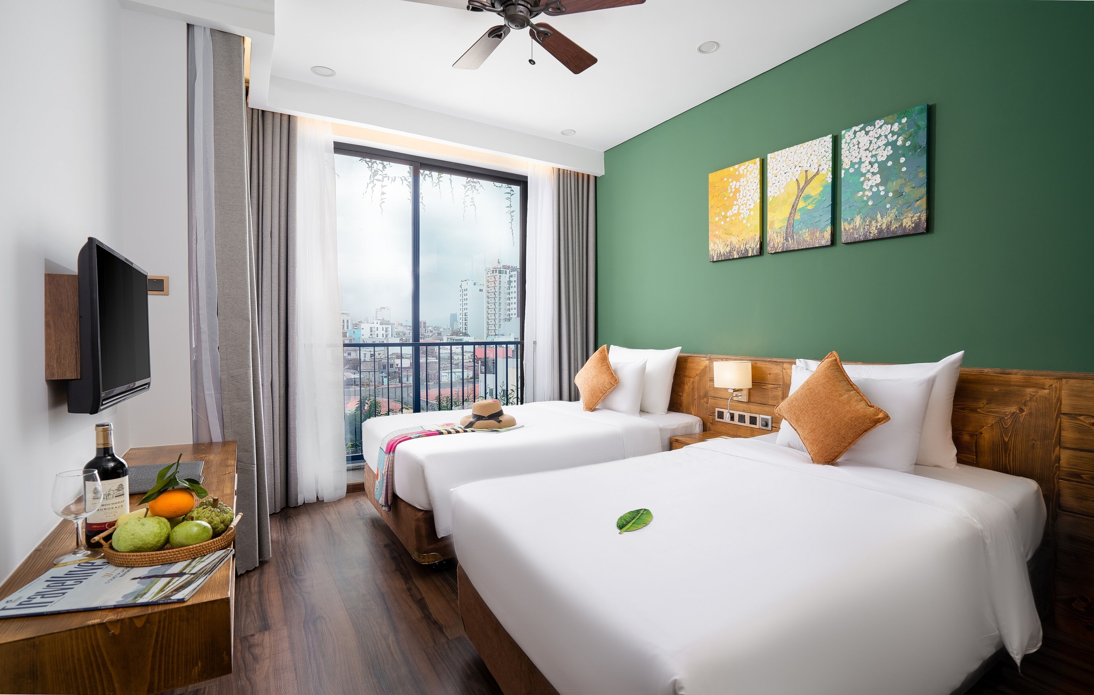 A hotel room with two beds, a television, a wooden desk with a bottle and fruit, and a city view.