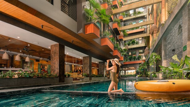 A modern indoor swimming pool with a central column, surrounded by balconies with plants, and a restaurant area in the background. - Chi House Danang (Da Nang)