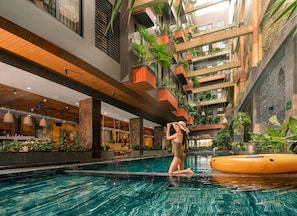 A modern indoor swimming pool with a central column, surrounded by balconies with plants, and a restaurant area in the background. - Chi House Danang (Da Nang)