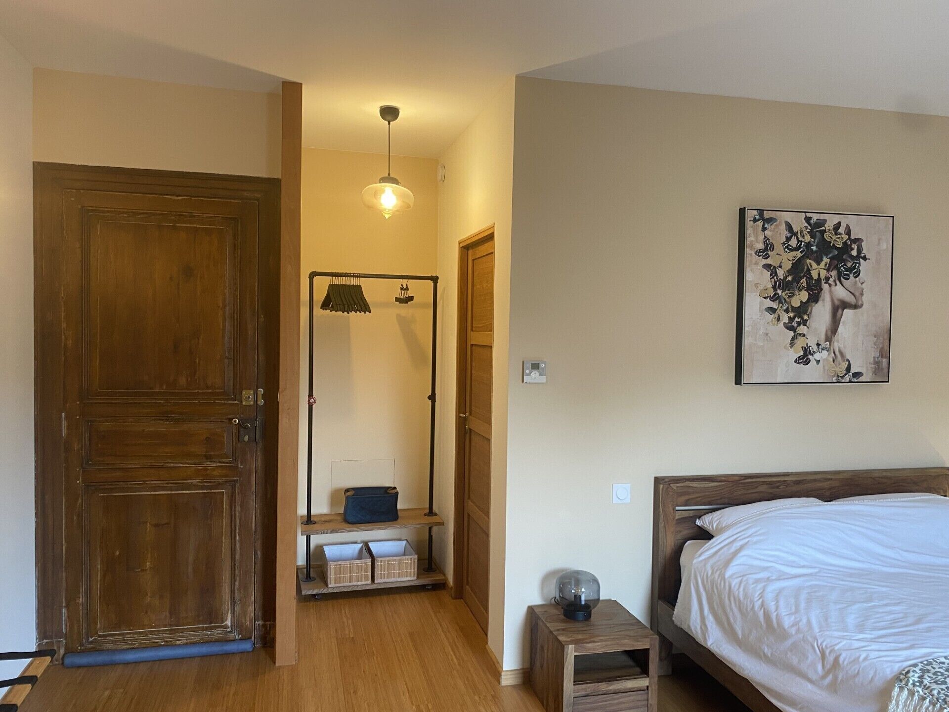 Une chambre avec une porte en bois, un lit avec une literie blanche, une petite table de chevet avec une lampe et une œuvre d’art accrochée au mur.