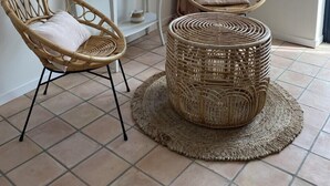 Una habitación con un espejo redondo, dos sillones de mimbre, una pequeña mesa consola blanca y un otoman redondo de mimbre.