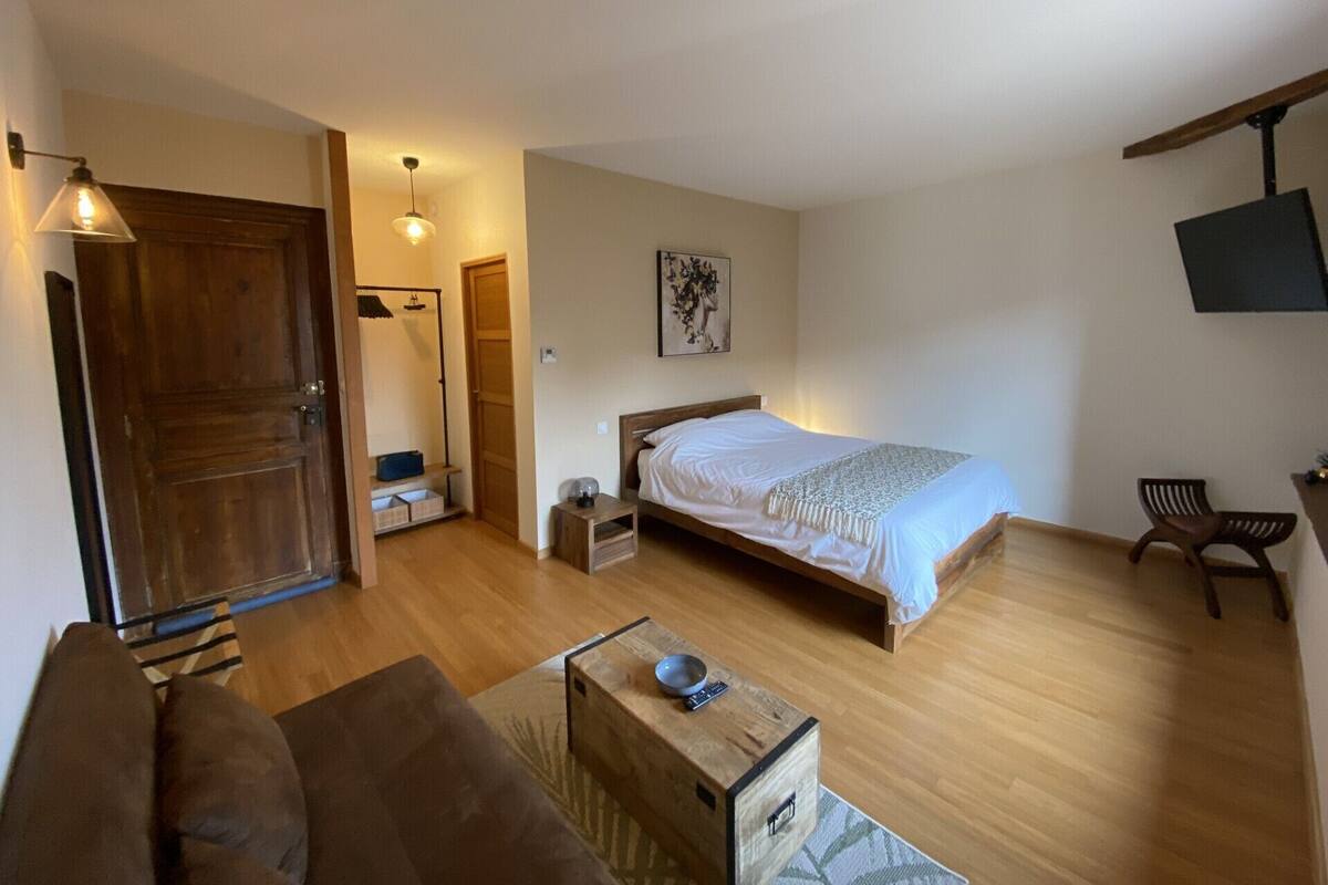 A bedroom with a wooden bed, a wooden floor, a wooden door, a small bedside table, a chair, a television mounted on the wall, and a framed picture on the wall.