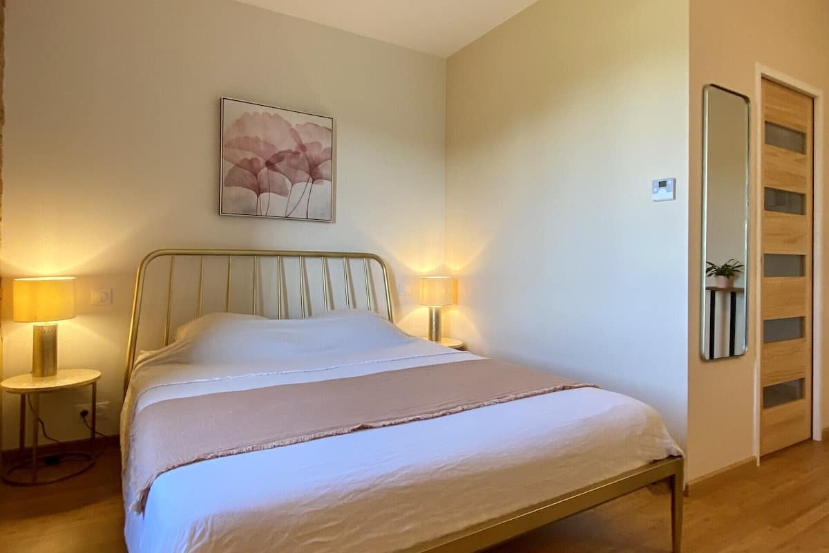 A neatly made bed in a room with a wooden floor, a bedside table with a lamp, and a wall-mounted mirror.