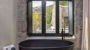A modern bathroom with a large freestanding bathtub, a stone wall, and a view of a pool and greenery outside.