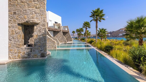 A pool area with stone walls and palm trees, leading to a view of the sea. - Aeonic Suites and Spa (Mykonos)