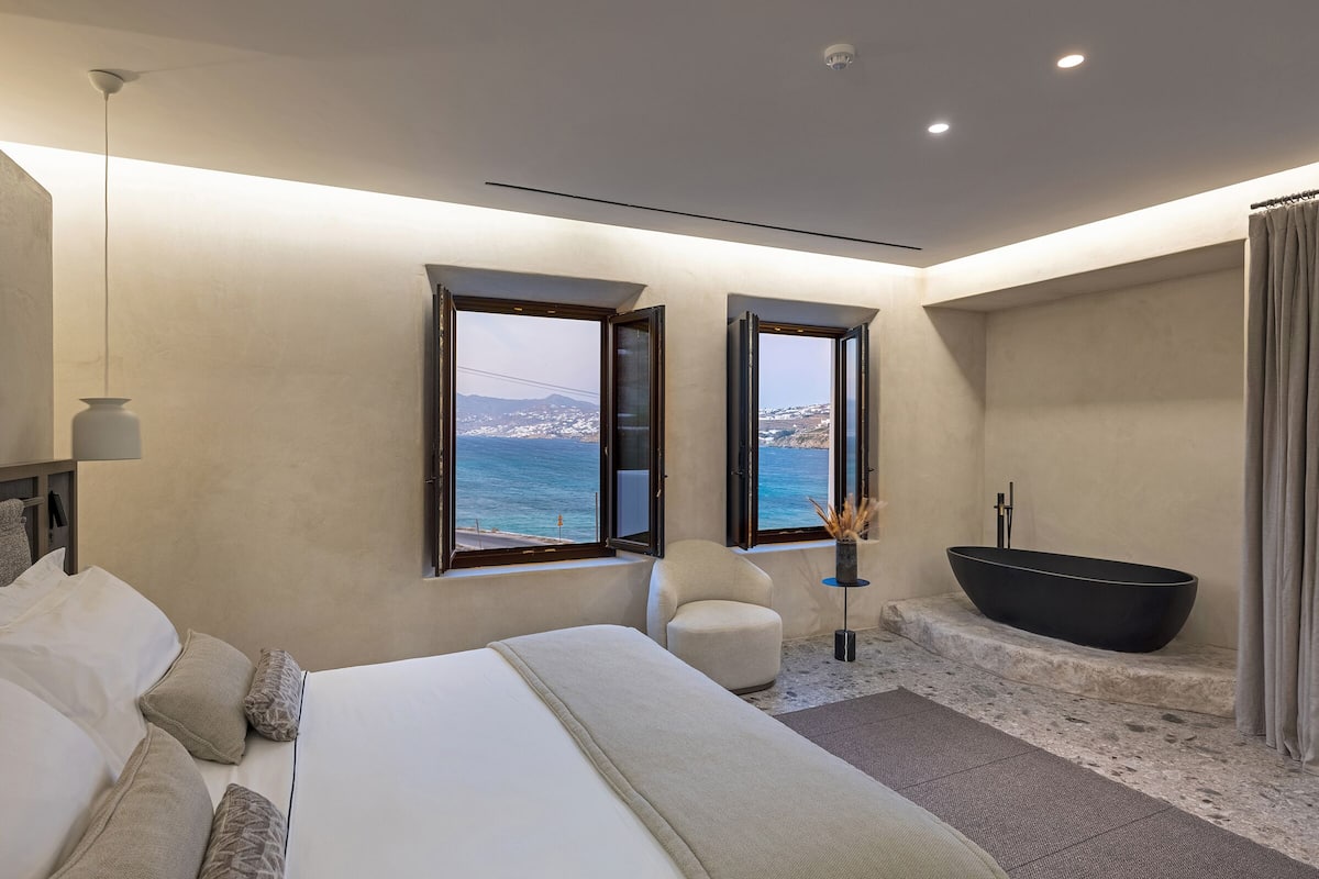 Un dormitorio moderno con una cama grande, una zona de estar, una bañera y vistas al mar.
