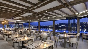 A restaurant with multiple tables and chairs, large windows offering a view of a cityscape and water at night. - Aeonic Suites and Spa (Mykonos)