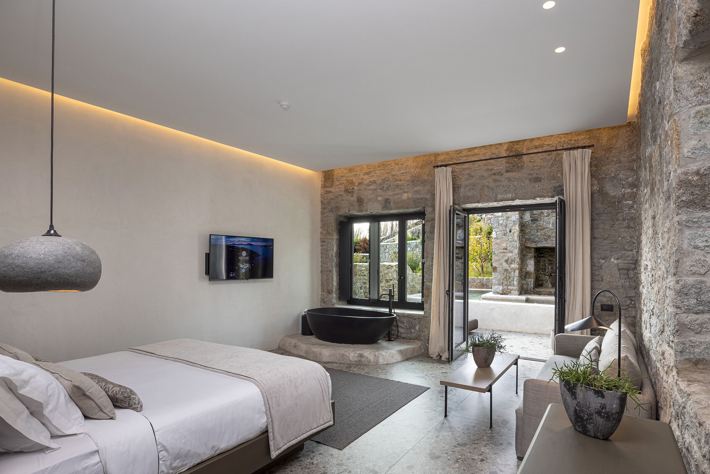 A modern bedroom with a large bed, a stone wall, a bathtub, and a view of the outdoors.