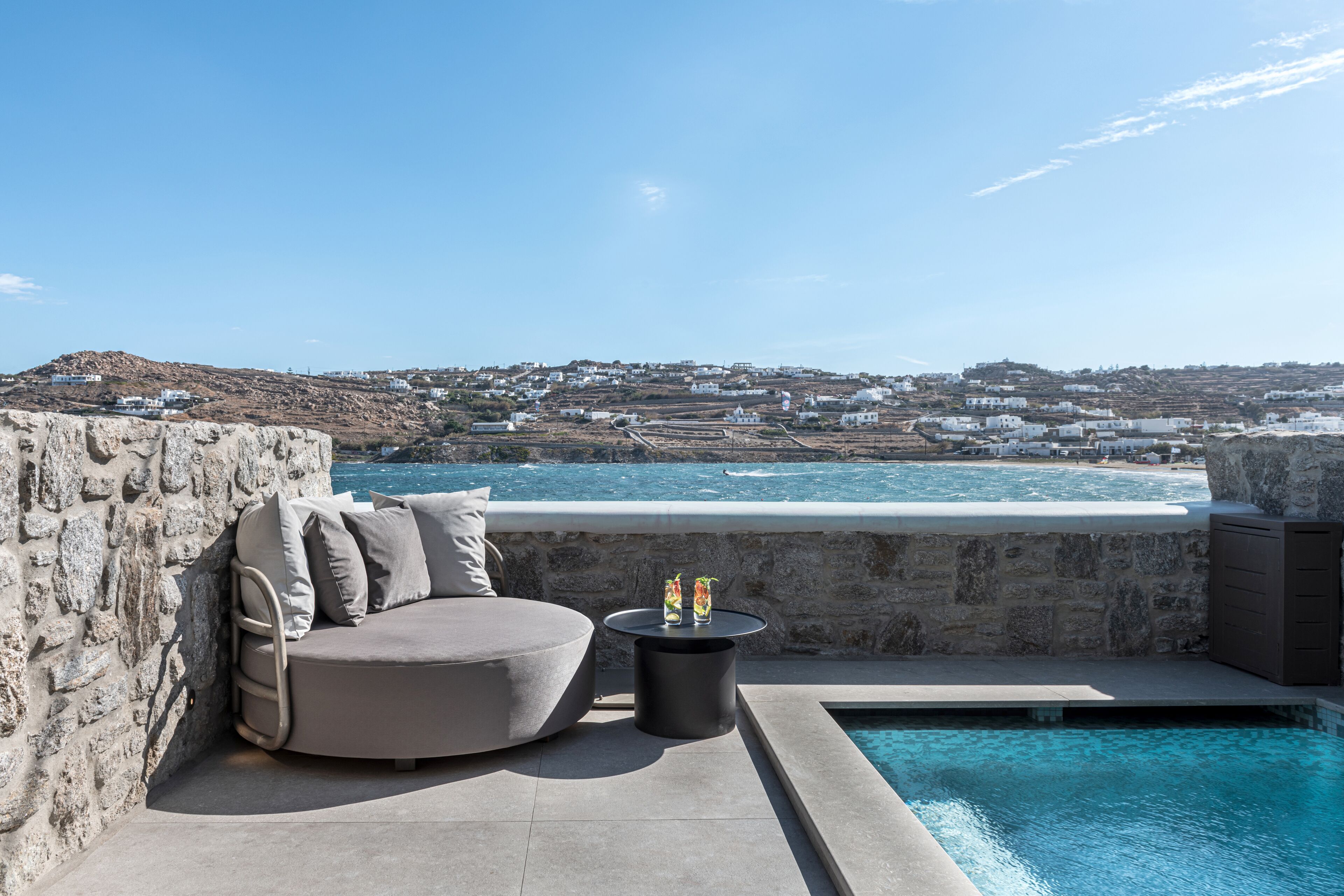 Un área junto a la piscina con un muro de piedra, una zona de estar con cojines y vistas a un pueblo costero.