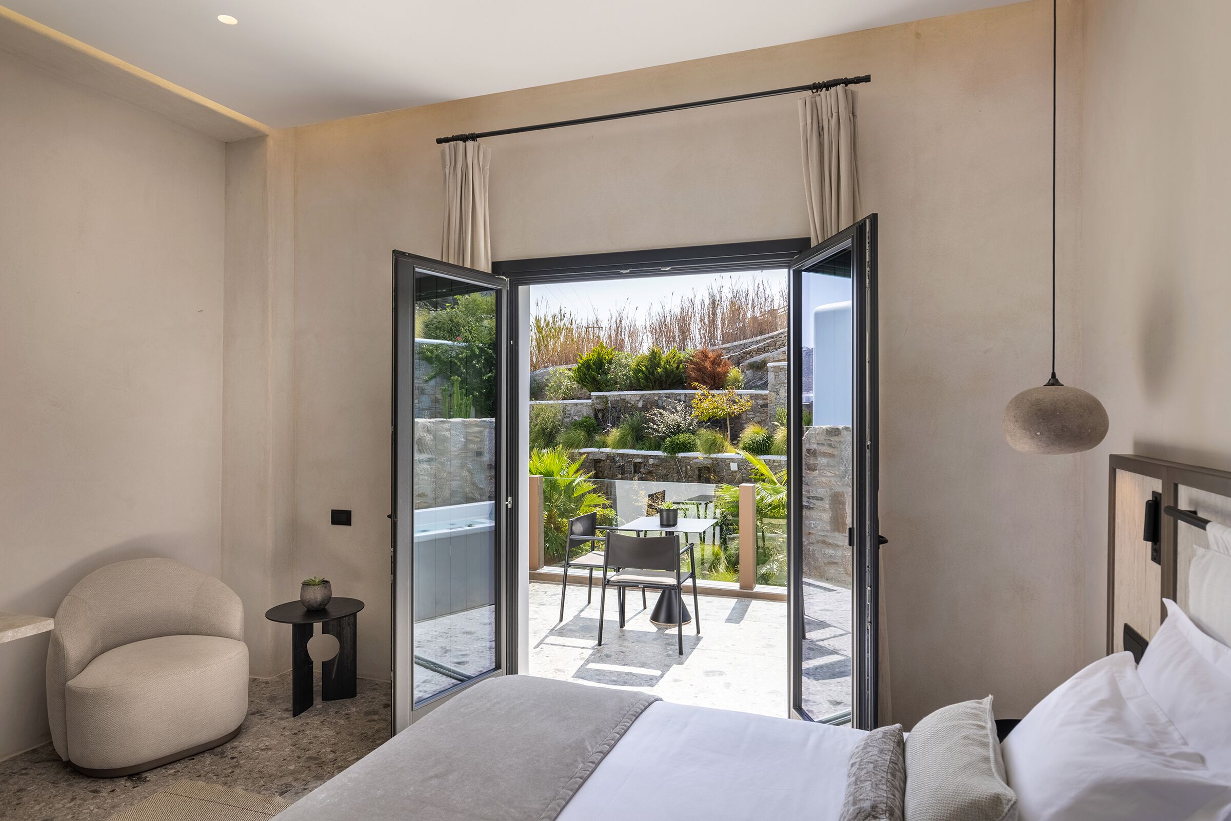 A modern bedroom with a large bed, a small round table, and a chair. There is a balcony with a table and chairs, and a view of a garden.