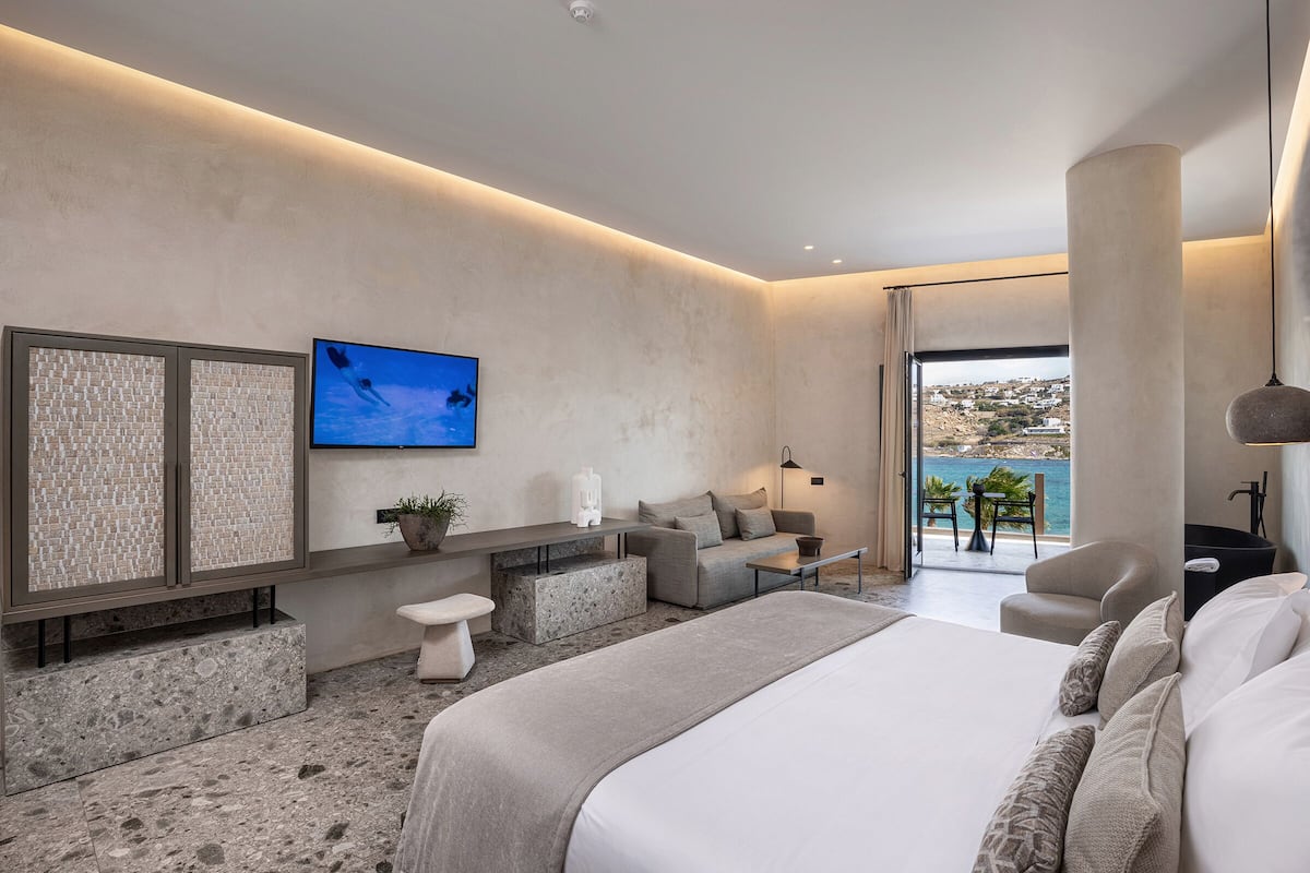 Una habitación de hotel moderna con una cama grande, un rincón de descanso con sofá y sillones, una mesita y un televisor en la pared.