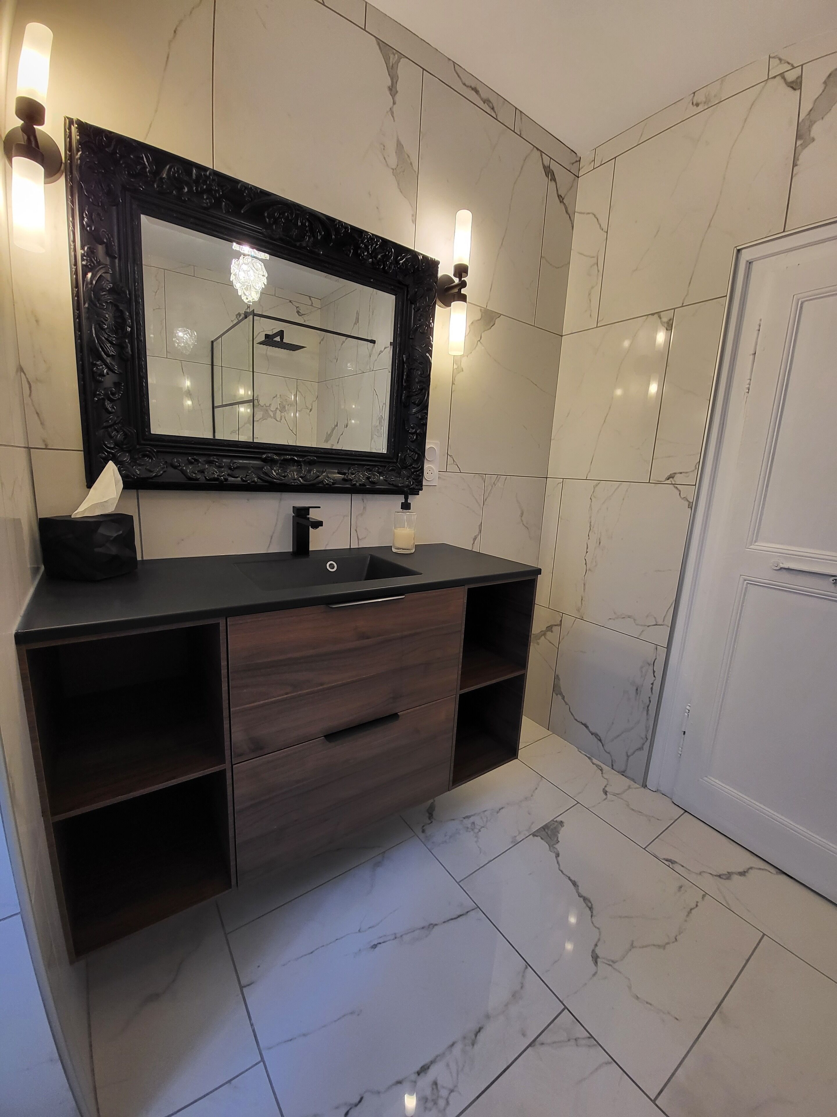 Un baño con suelo de mármol, un mueble de tocador de madera oscura con un espejo de marco negro y dos luces empotradas.