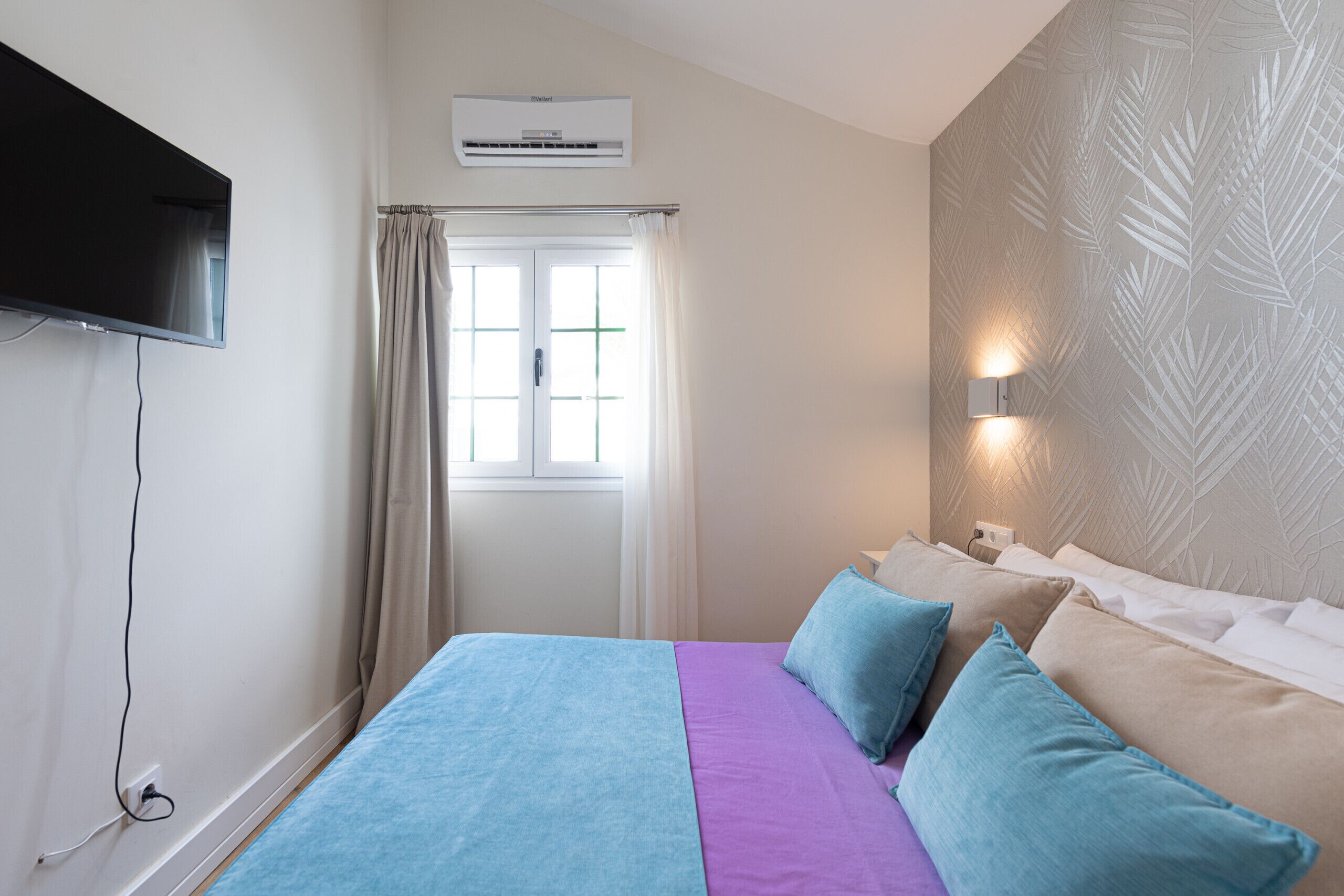 A bedroom with a bed, a TV, an air conditioner, a window with curtains, and a wall with a leaf pattern.
