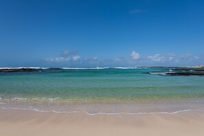 Een strand met helder turquoise water, een zandstrand en veraf gelegen rotsformaties.