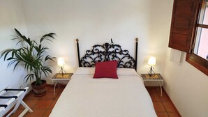 A bed with a decorative headboard, two bedside tables with lamps, a potted plant, and a window with wooden shutters. - Casa Montecote Eco Resort - Apartment Romero (VEJER)