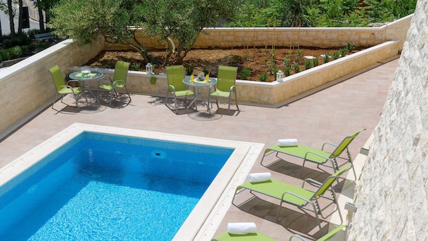 A swimming pool with green lounge chairs and a table with chairs, surrounded by a stone wall and greenery.