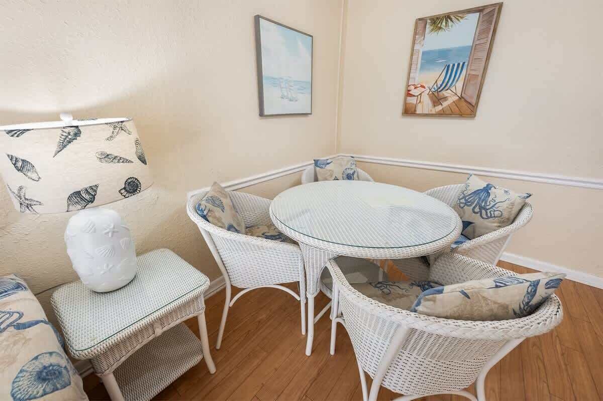 Une pièce d’angle avec une table ronde en verre et des chaises en osier blanc, un petit tabouret blanc et un mur orné de tableaux encadrés.