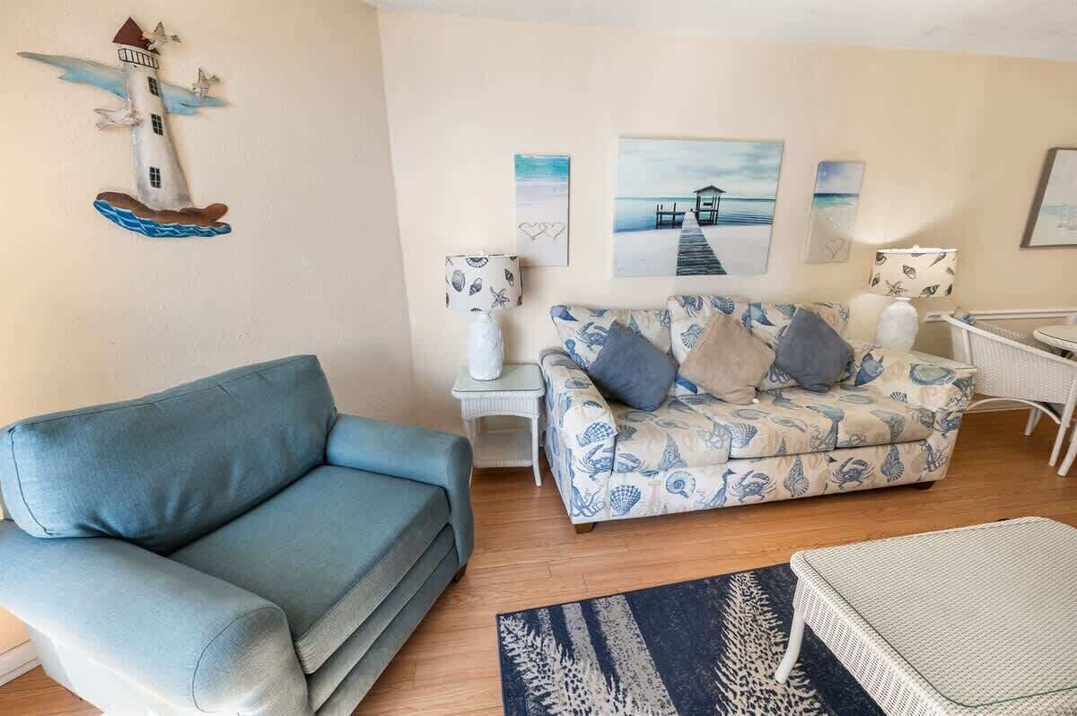 A living room with a blue sofa, a patterned sofa, a small table, and a chair. There are wall art pieces and a decorative wall hanging.