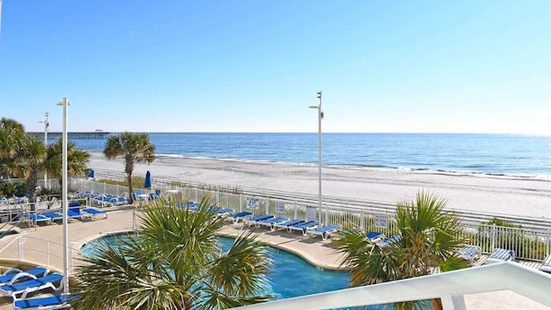 A pool area with lounge chairs and palm trees, overlooking a beach and ocean.
