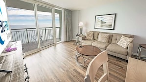 A spacious living room with a large sliding glass door leading to a balcony, a beige sofa, a round wooden coffee table, and a framed picture on the wall.
