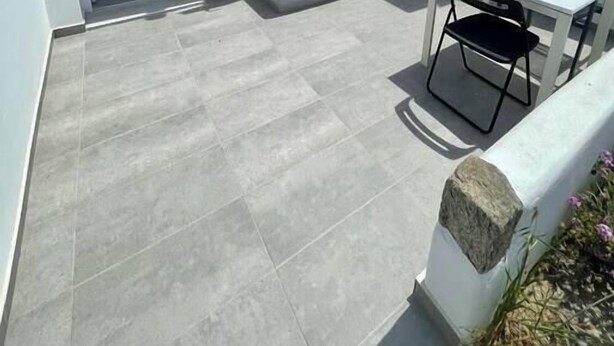 En hvid bygning med fliseterrasse, et lille bord med stol og et vindue med skodder.
