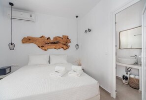 Ein Schlafzimmer mit einem großen Bett, weißer Bettwäsche, einem Holzbild an der Wand, einem Nachttisch, einer Wandlampe und einem Badezimmer mit Waschbecken und Spiegel.