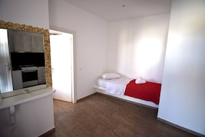 A bedroom with a bed, a red blanket, a white pillow, a white nightstand, and a kitchen area with cabinets and a stove.