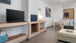 En moderne stue med et fladskærms-tv, en pejs, et spiseområde med bord og stole, og et køkken der kan ses gennem et åbent vindue.