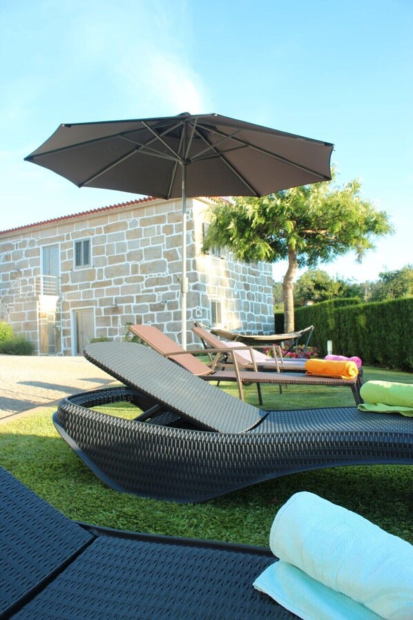 A stone house with a patio area featuring two lounge chairs, a large umbrella, and a small pool.
