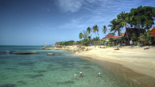 Una playa con agua turquesa cristalina, palmeras y casas de playa con tejados rojos.