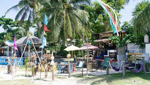 Un café junto a la playa con terraza al aire libre, palmeras y un cartel anunciando bebidas y comida.