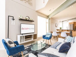 Un salon moderne avec un téléviseur à écran plat, un canapé, une table basse et un coin repas comprenant des chaises, avec une cuisine en arrière-plan.