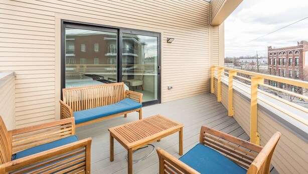 A balcony with wooden furniture, a glass door, and a view of buildings. - Upscale 3BR Penthouse by CozySuites (Indianapolis)