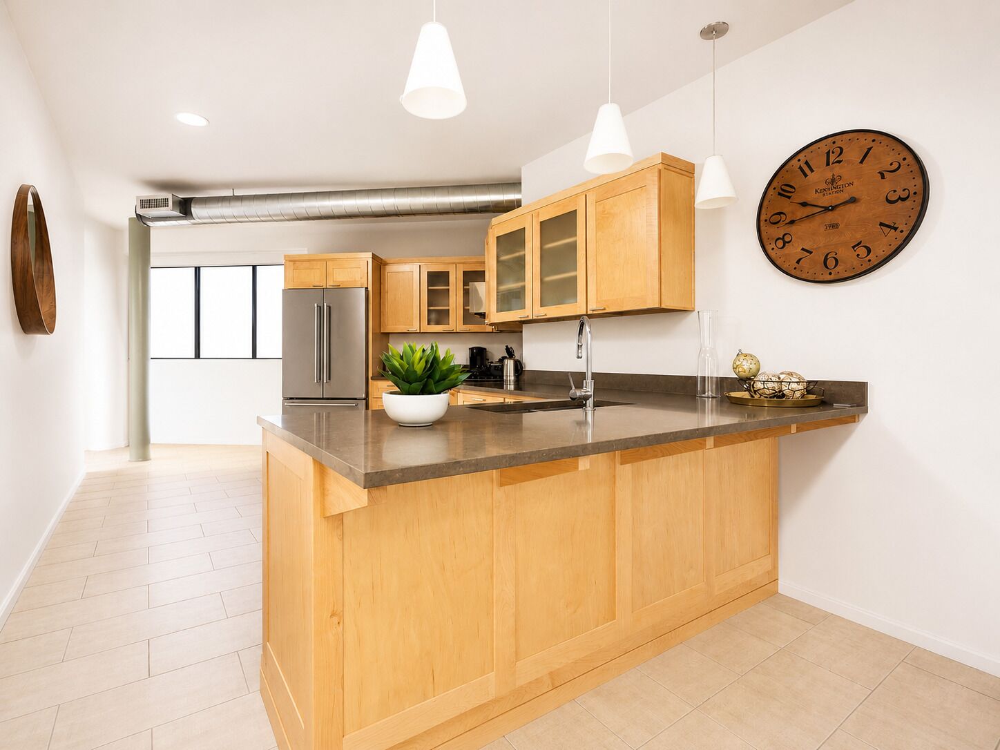 Een moderne keuken met houten kastjes, een roestvrijstalen koelkast, een groot eiland met een spoelbak, hanglampen en een wandklok.