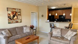 A living room with two sofas, a wooden coffee table, and a kitchen area with wooden cabinets and pendant lights.