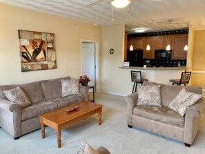 A living room with two sofas, a wooden coffee table, and a kitchen area with wooden cabinets and pendant lights.