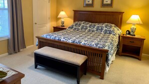 A bedroom with a wooden bed, two bedside tables with lamps, a bench, a door, and two framed pictures on the wall.
