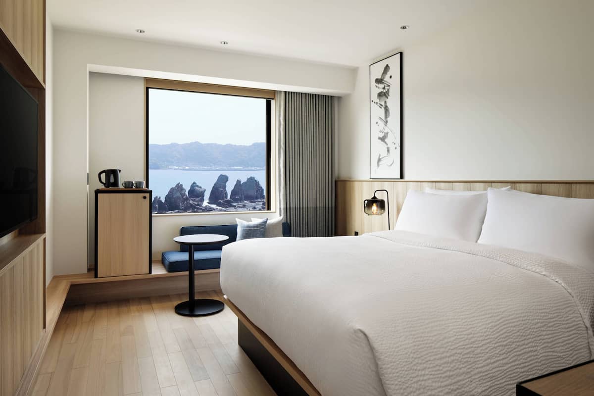 A modern hotel room with a large bed, a small seating area, a view of the sea, and a flat-screen TV.