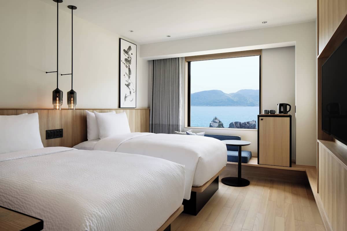 A modern hotel room with two beds, a large window with a view of the sea, a small table, and a television.