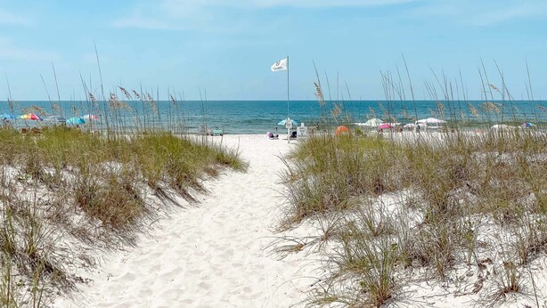 Un sentier de sable qui longe la plage et mène à la mer, avec des parasols de plage et un drapeau.