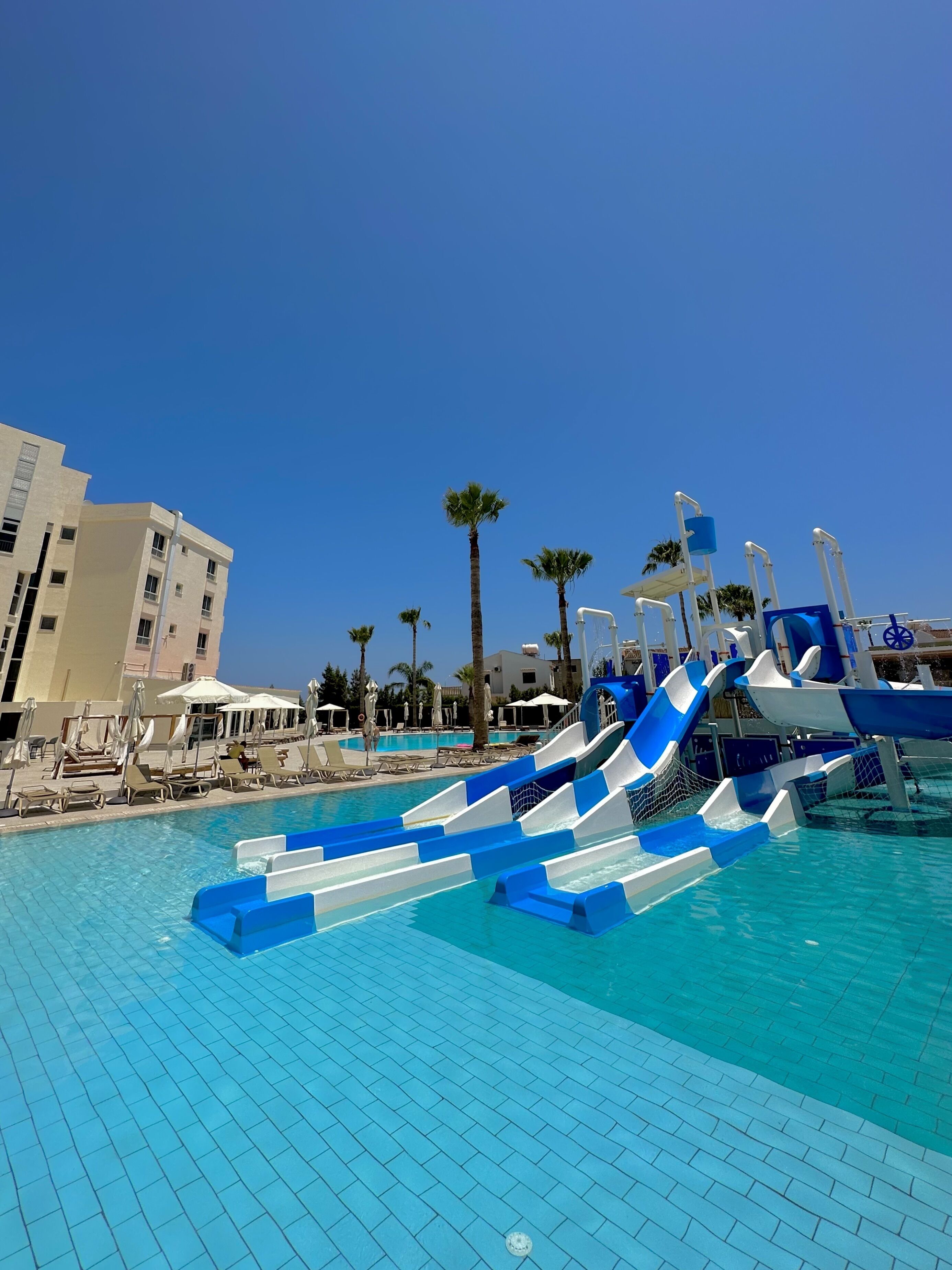 A swimming pool with multiple slides, lounge chairs, umbrellas, and palm trees.