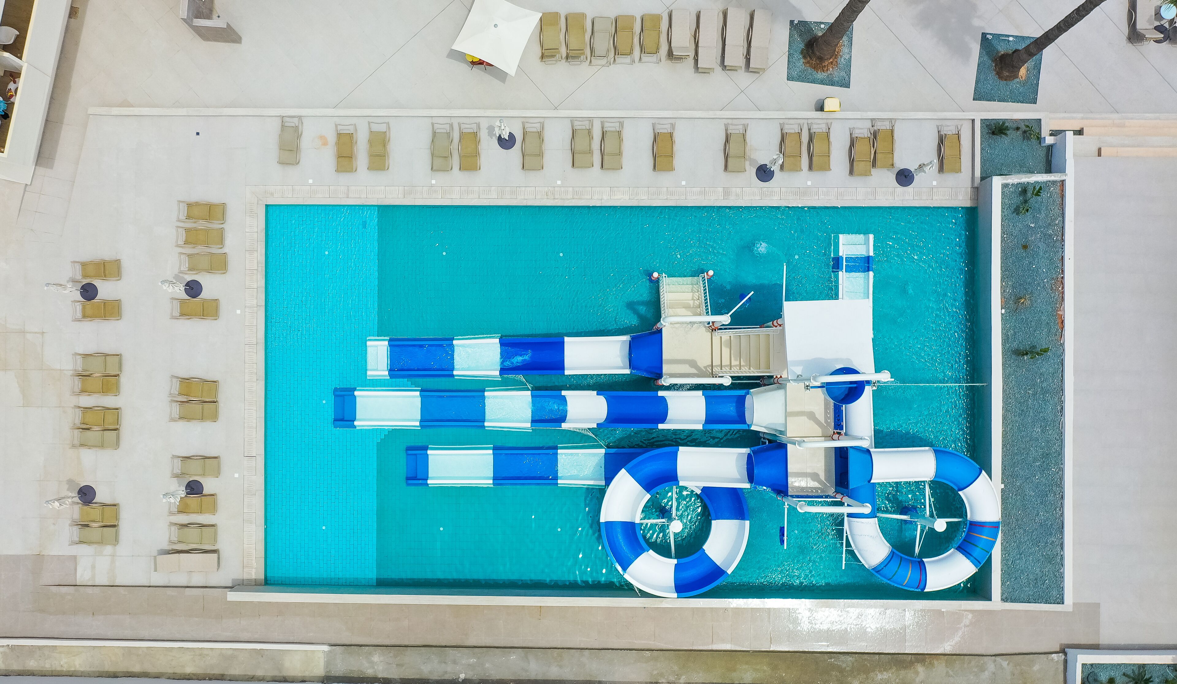 Vista aérea de una zona de piscina con múltiples sillas de descanso, un tobogán acuático y un flotador salvavidas.