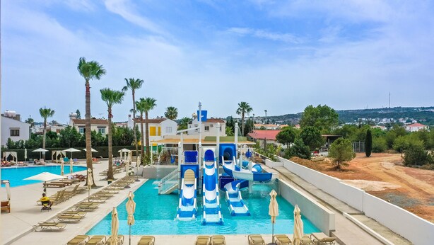 A swimming pool with lounge chairs and palm trees, a water slide, and a view of the surrounding area. - Toxotis Hotel  (Paralimni)